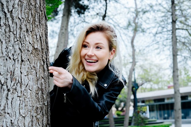 Junge überraschte Frau, die sich hinter dem Baum versteckt und mit dem Finger in einem Park mit schwarzer Lederjacke darauf hinweist