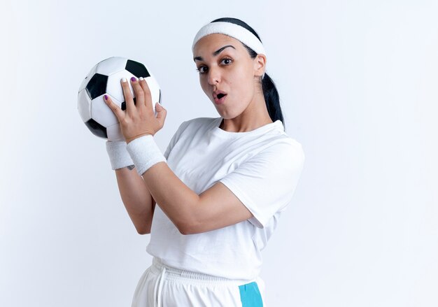 Junge überraschte kaukasische sportliche Frau, die Stirnband und Armbänder trägt, hält Ball lokalisiert auf weißem Raum mit Kopienraum