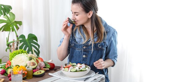 Junge und glückliche Frau, die Salat am Tisch isst, auf einem hellen Hintergrund in Jeanskleidung. Das Konzept eines gesunden hausgemachten Lebensmittels.