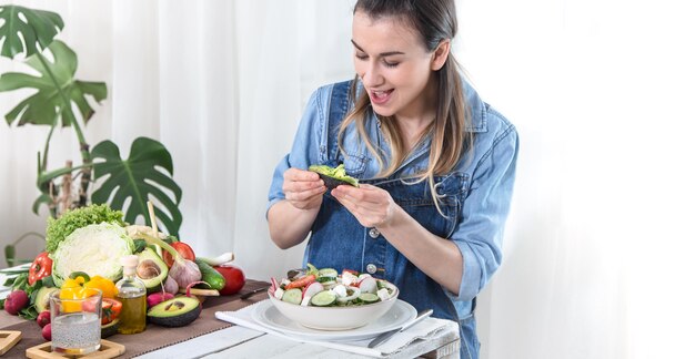 Junge und glückliche Frau, die Salat mit Bio-Gemüse am Tisch auf hellem Hintergrund in Jeanskleidern isst. Das Konzept eines gesunden hausgemachten Lebensmittels.