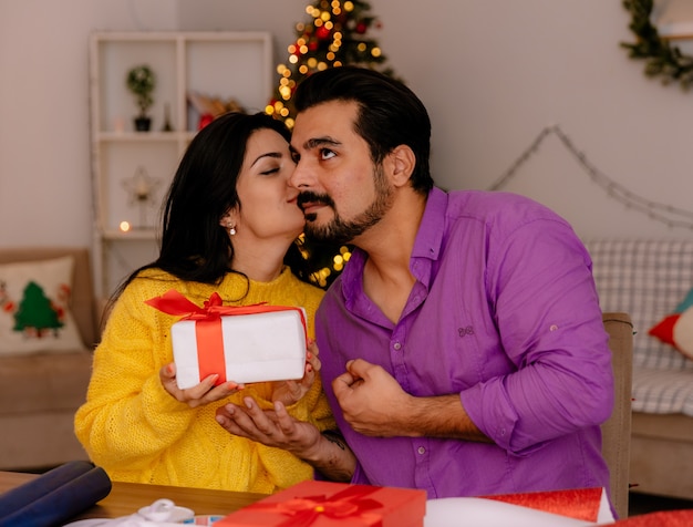 junge und schöne Paarfrau, die ein Geschenk homding und ihren Freund im weihnachtlich geschmückten Raum mit Weihnachtsbaum im Hintergrund küsst