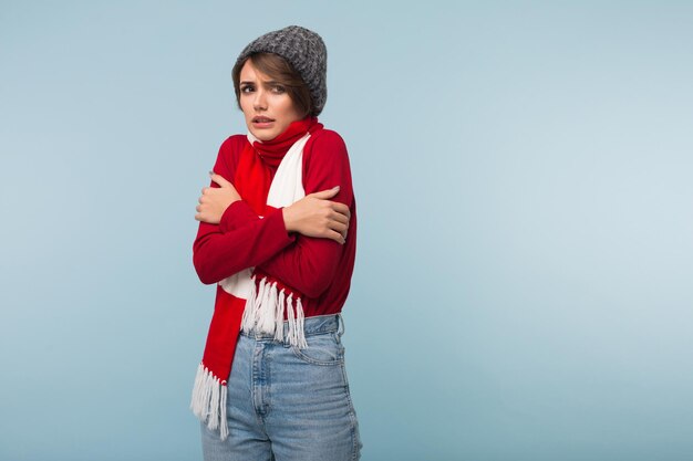 Junge ungesunde Frau mit dunklen kurzen Haaren in rotem Pullover, Schal und Strickmütze, die vor Kälte zittert, während sie traurig über blauem Hintergrund zur Seite schaut