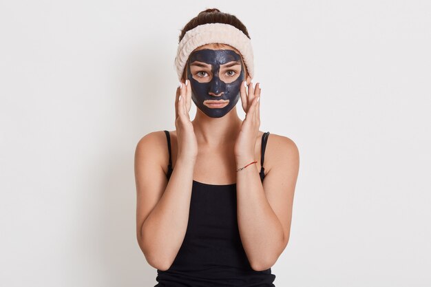 Junge unglückliche Hausfrau mit schwarzer kosmetischer Maske auf Gesicht, das isoliert über weißer Wand steht und ihre Wangen mit den Fingern berührt