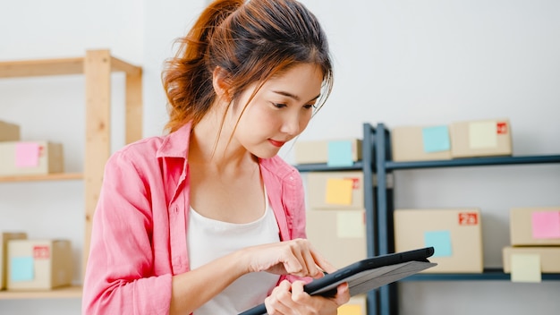 Junge Unternehmerin aus Asien prüft die Produktbestellung auf Lager und speichert sie auf dem Tablet-Computer zu Hause. Kleinunternehmer, Online-Marktzustellung, freiberufliches Lifestyle-Konzept.
