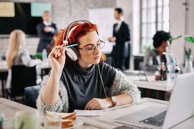 Junge Unternehmerin, die einen Laptop verwendet und über E-Mails nachdenkt, die sie erhalten hat, während sie bei der Arbeit Musik über Kopfhörer hört. Es gibt Menschen im Hintergrund