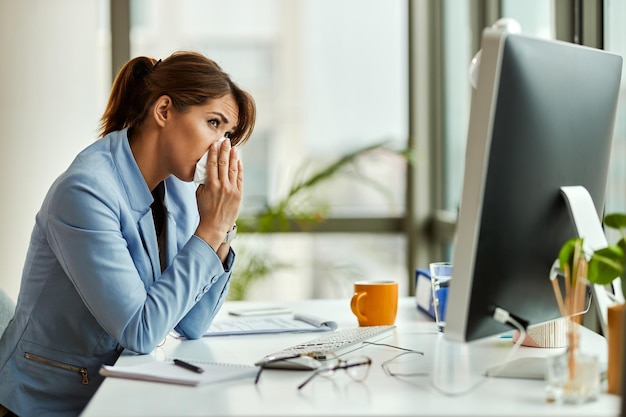 Junge Unternehmerin niest in ein Taschentuch, während sie an ihrem Schreibtisch arbeitet