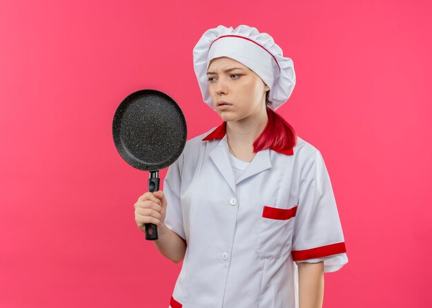 Junge verärgerte blonde Köchin in Kochuniform hält Bratpfanne und schaut zur Seite isoliert auf rosa Wand