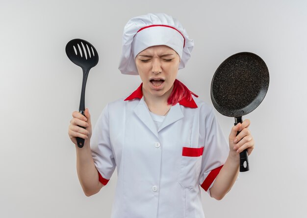 Junge verärgerte blonde Köchin in Kochuniform hält Bratpfanne und Spatel isoliert auf weißer Wand