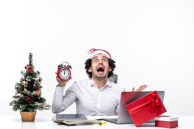 Junge verärgerte Geschäftsperson mit Weihnachtsmannhut und Halten der Uhr und des Sitzens im Büro auf dunklem Hintergrund