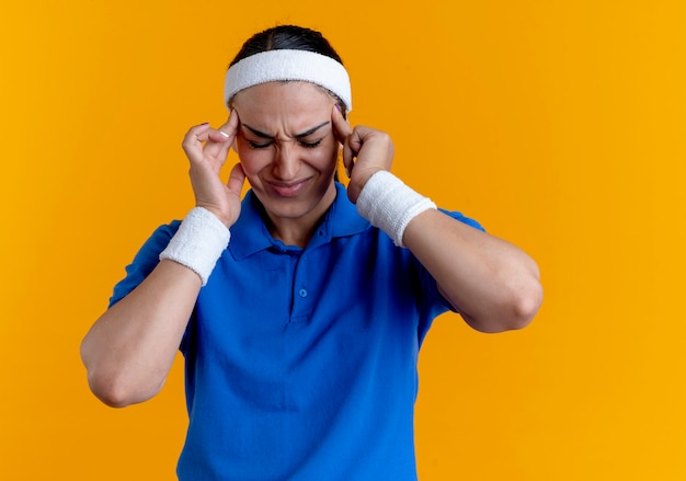 Junge verärgerte kaukasische sportliche Frau, die Stirnband und Armbänder trägt, hält Kopf mit geschlossenen Augen auf Orange mit Kopienraum