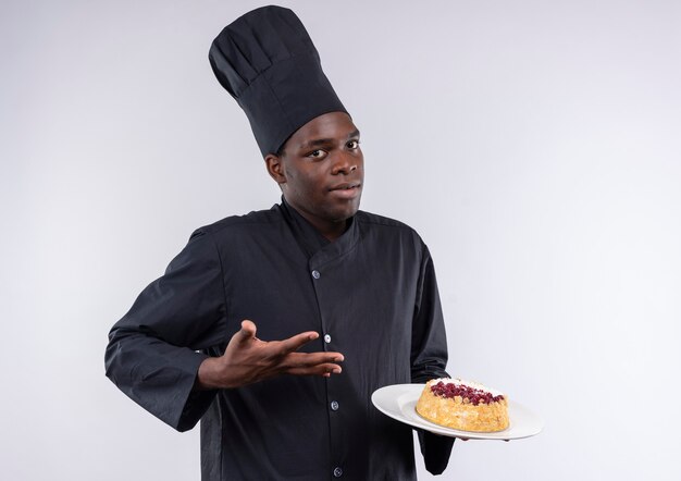Junge verwirrte afroamerikanische Köchin in der Kochuniform hält und zeigt auf Kuchen auf Teller auf Weiß mit Kopienraum