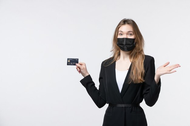Junge verwirrte Unternehmerin im Anzug, die ihre medizinische Maske trägt und eine Bankkarte an der weißen Wand hält