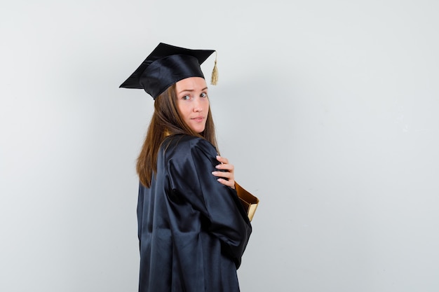 Junge weibliche Absolventin, die beim Betrachten der Kamera im akademischen Kleid aufwirft und glamourös schaut. Vorderansicht.