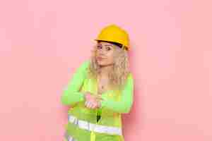 Kostenloses Foto junge weibliche baumeisterin der vorderansicht im gelben helm des grünen bauanzugs, der gerade auf dem rosa raumjobarchitektur-konstruktionsfoto aufwirft