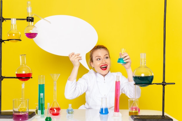 Junge weibliche Chemikerin der Vorderansicht, die im weißen Anzug mit verschiedenen Lösungen sitzt und weißes Zeichen hält, das auf dem gelben Boden Chemie Wissenschaft s lächelt