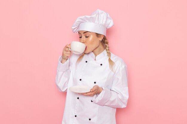 Junge weibliche Köchin der Vorderansicht im weißen Kochanzug, der heißen Kaffee auf dem rosa Raumkoch trinkt