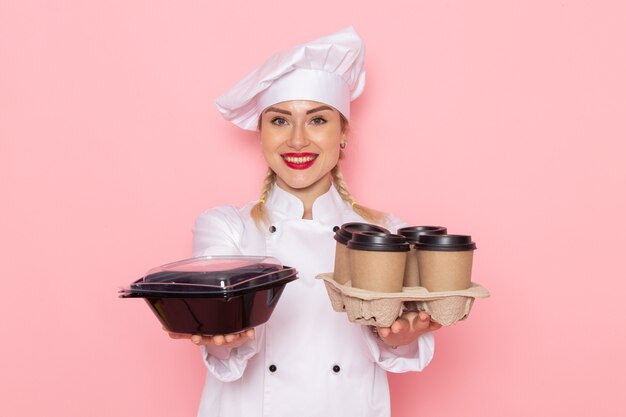 Junge weibliche Köchin der Vorderansicht im weißen Kochanzug, der Plastikkaffeetassen und Lebensmittelverpackung auf dem rosa Raumkochfoto hält