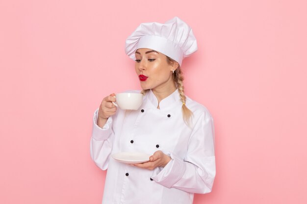 Junge weibliche Köchin der Vorderansicht im weißen Kochanzug, der weiße Tasse mit Kaffee auf dem rosa Raumkoch hält