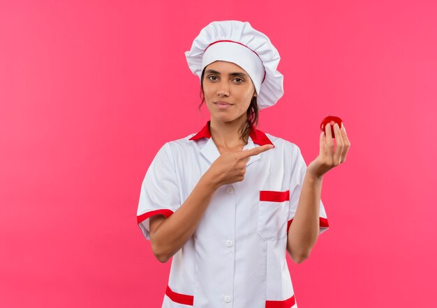 junge weibliche Köchin, die Kochuniform hält und Tomate auf isolierte rosa Wand mit Kopienraum zeigt