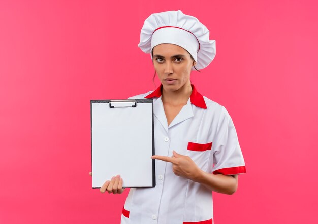 junge weibliche Köchin, die Kochuniform hält und zeigt auf Zwischenablage auf isolierter rosa Wand mit Kopienraum