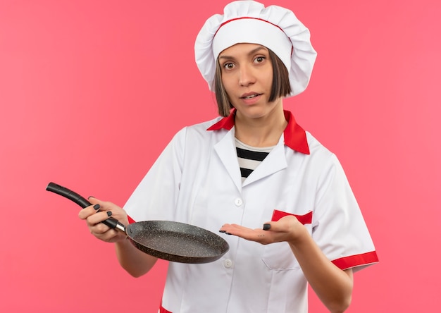 Junge weibliche Köchin in der Kochuniform, die mit der Hand an der Bratpfanne hält und Kamera auf rosa Hintergrund lokalisiert hält