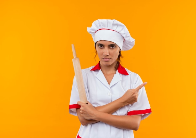 junge weibliche Köchin trägt Kochuniform, die Handa hält, die Nudelholz hält und zur Seite auf isolierte gelbe Wand mit Kopienraum zeigt