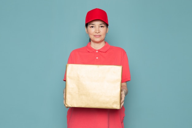 Junge weibliche kurier in rosa t-shirt rote kappe, die box an der blauen wand hält