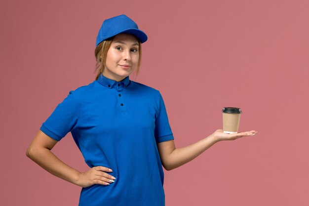 Junge weibliche Kurierin der Vorderansicht in der blauen Uniform, die das Halten der Lieferschale des Kaffees auf der rosa Wanddienstjobuniformlieferungsfrau aufwirft