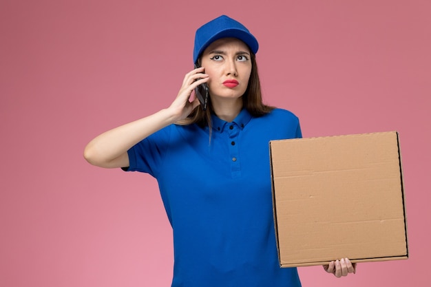 Junge weibliche Kurierin der Vorderansicht in der blauen Uniform und im Umhang, die Nahrungsmittelbox hält, die am Telefon an der rosa Wand spricht