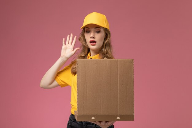 Kostenloses Foto junge weibliche kurierin der vorderansicht in der gelben uniform, die das liefernahrungsmittelpaket hält, das es auf rosa schreibtischjobuniformlieferdienstarbeiter betrachtet