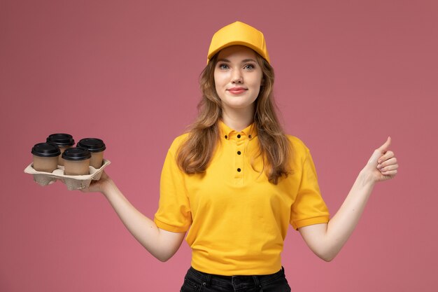 Junge weibliche Kurierin der Vorderansicht in der gelben Uniform, die Kaffeetassen hält und auf dem rosa Hintergrundschreibtischjobuniformlieferdienstarbeiter lächelt