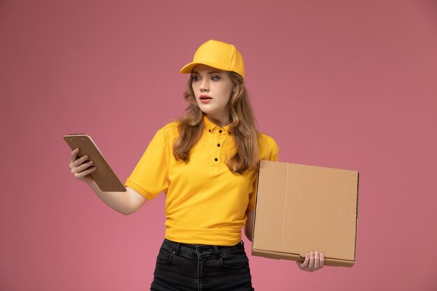 Junge weibliche Kurierin der Vorderansicht in der gelben Uniform, die Paket mit Essen und Notizblock auf dem rosa Hintergrundschreibtischjobuniformlieferdienstarbeiter hält