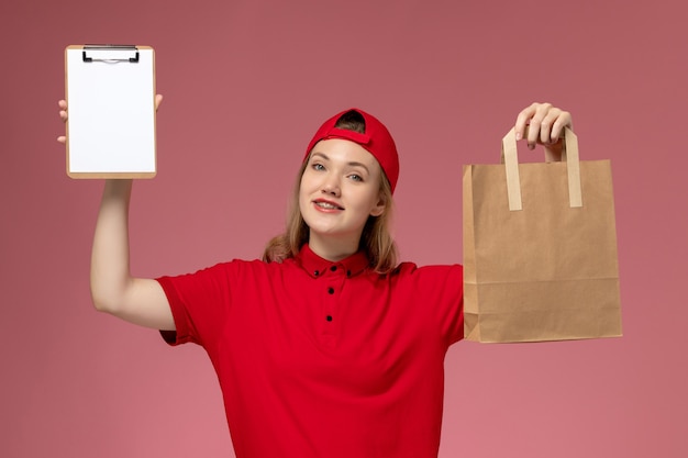 Junge weibliche Kurierin der Vorderansicht in der roten Uniform, die das Liefernahrungsmittelpaket und den Notizblock hält, der auf hellrosa Wand lächelt