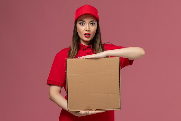 Junge weibliche Kurierin der Vorderansicht in der roten Uniform, die die Liefernahrungsmittelbox auf dem rosa Schreibtischdienstlieferungsuniformunternehmen hält