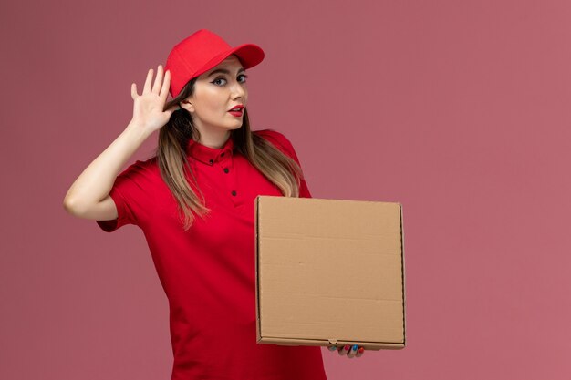 Junge weibliche Kurierin der Vorderansicht in der roten Uniform, die die Liefernahrungsmittelbox hält, die versucht, auf der rosa Hintergrunddienstlieferungsuniformfirma zu hören