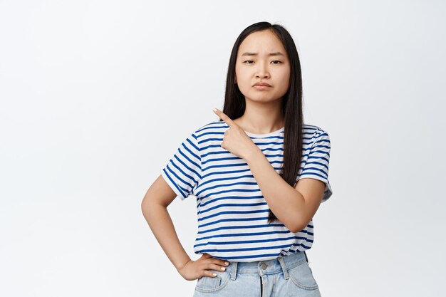 Junge, widerstrebende asiatische Frau, die mit dem Finger auf die obere linke Ecke zeigt und das Gesicht verzieht, enttäuscht, dass sie unzufrieden ist, weil etw auf weißem Hintergrund steht