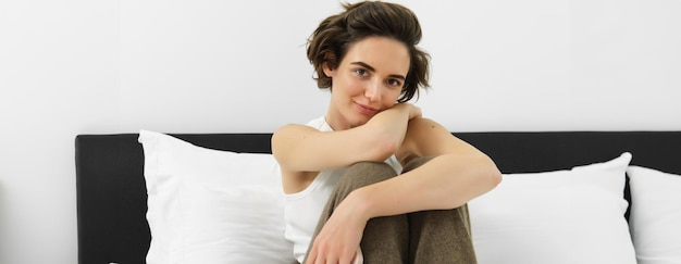 Kostenloses Foto junge zärtliche frau in ihrem gemütlichen pyjama sitzt auf dem bett im schlafzimmer und lächelt mit einem sinnlichen lächeln
