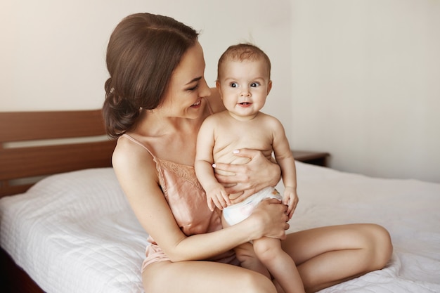 Junge zarte glückliche Mutter, die ihr neugeborenes Baby lächelnd am Morgen auf dem Bett sitzend umarmt.