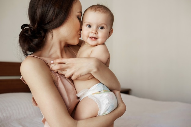 Junge zarte glückliche Mutter, die ihr neugeborenes Baby lächelnd am Morgen auf dem Bett sitzend umarmt.