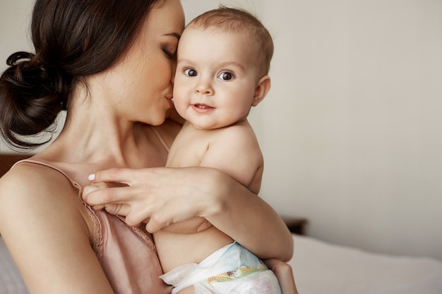 Junge zarte glückliche Mutter, die ihr neugeborenes Baby lächelnd am Morgen auf dem Bett sitzend umarmt