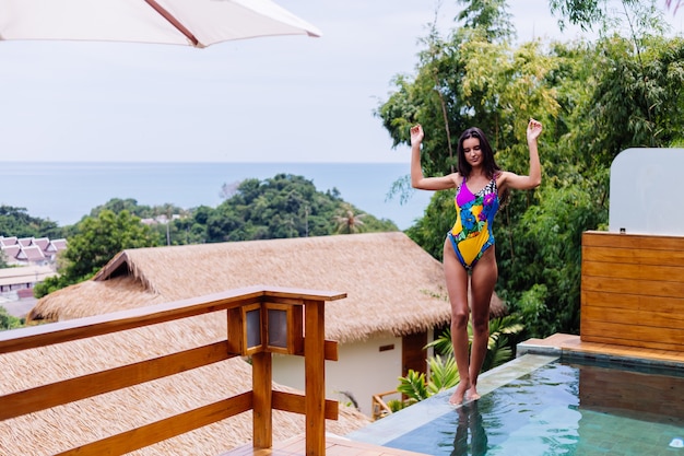 Junge ziemlich positive glückliche schlanke Frau im bunten Badeanzug an der Luxusvilla des erstaunlichen Hotels, das schönen Tag im Urlaub in Thailand genießt
