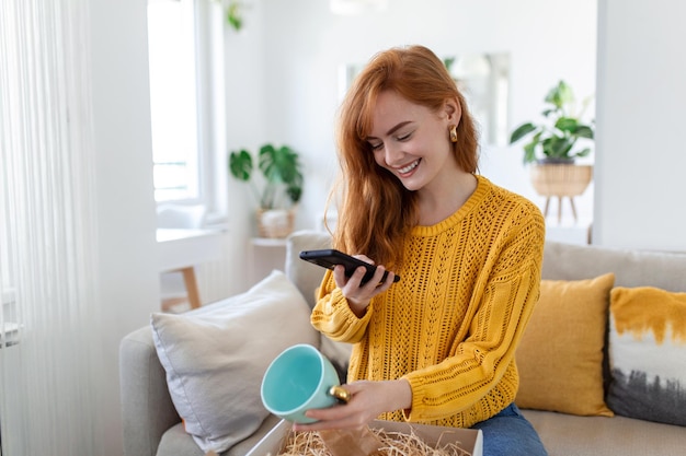 Junge zufriedene, glückliche Shopaholic-Kundin sitzt auf dem Sofa und packt das Online-Shopping-Versandkonzept der Paketzustellbox aus, das Fotos des Produkts macht, um es in den sozialen Medien zu veröffentlichen