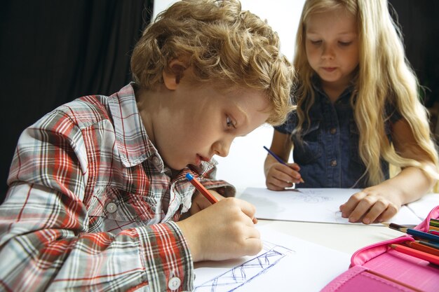 Jungen und Mädchen bereiten sich nach einer langen Sommerpause auf die Schule vor. Zurück zur Schule. Kleine kaukasische Modelle, die zusammen auf weißem und schwarzem Hintergrund zeichnen. Kindheits-, Bildungs-, Ferien- oder Hausaufgabenkonzept.