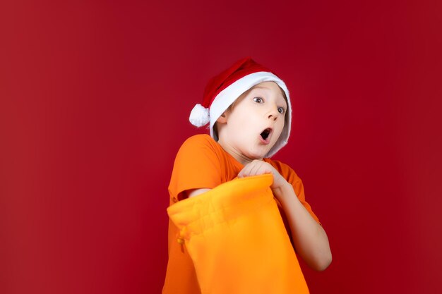 Jungen weihnachtsmütze von santa claus, die vor einem roten hintergrund steht, steckte seine hand in gelbe geschenktüten