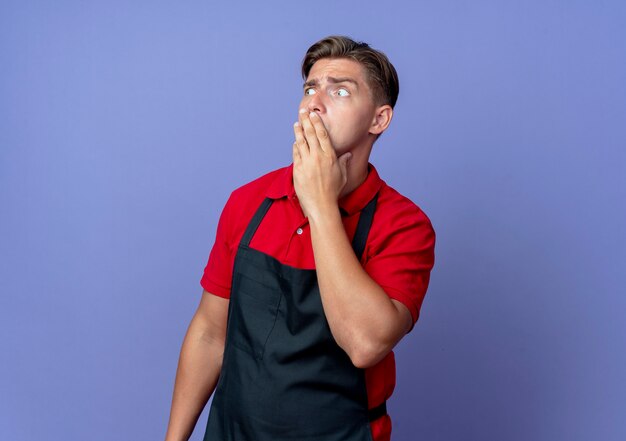 Junger ängstlicher blonder männlicher Friseur in der Uniform legt Hand auf Mund, der Seite betrachtet, die auf violettem Raum mit Kopierraum isoliert ist