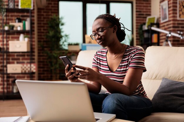 Junger afrikanisch-amerikanischer Freiberufler hält Smartphone, tippt Nachricht ein und chattet im sozialen Netzwerk. Fernarbeiter, die Pause machen, mit dem Handy im Internet surfen, auf der Couch sitzen
