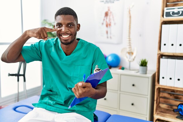 Junger afrikanisch-amerikanischer Mann, der in einer Schmerzklinik arbeitet und mit dem Finger auf Gesicht und Nase zeigt, lächelt fröhliches Schönheitskonzept