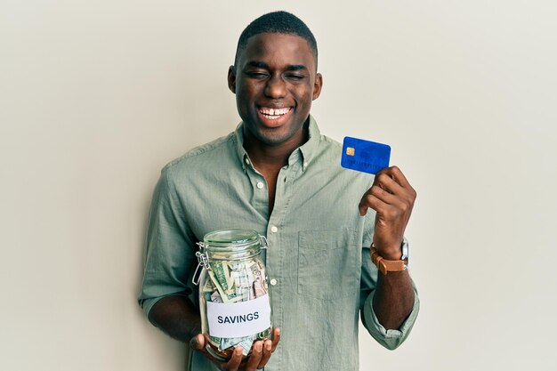 Junger afrikanisch-amerikanischer Mann, der Kreditkarte und Glas mit Dollars hält, lächelt und lacht laut, weil lustiger verrückter Witz.