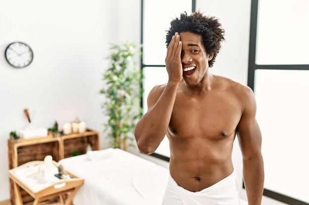 Junger afrikanisch-amerikanischer Mann mit nacktem Oberkörper und Handtuch, der im Schönheitszentrum steht und ein Auge mit der Hand bedeckt, selbstbewusstes Lächeln im Gesicht und überraschende Emotionen