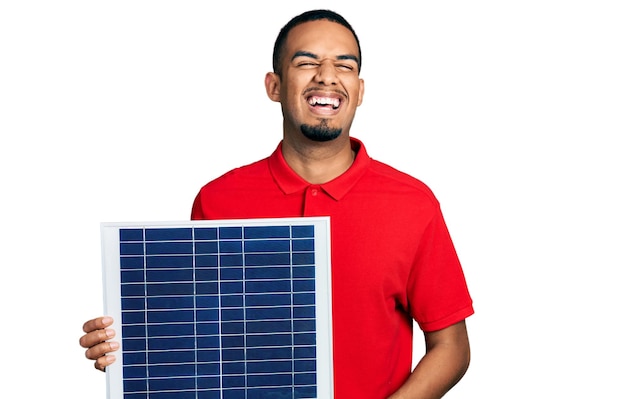Junger afrikanisch-amerikanischer Mann mit Photovoltaik-Solarpanel feiert verrückt und erstaunt über den Erfolg mit offenen Augen, die aufgeregt schreien.
